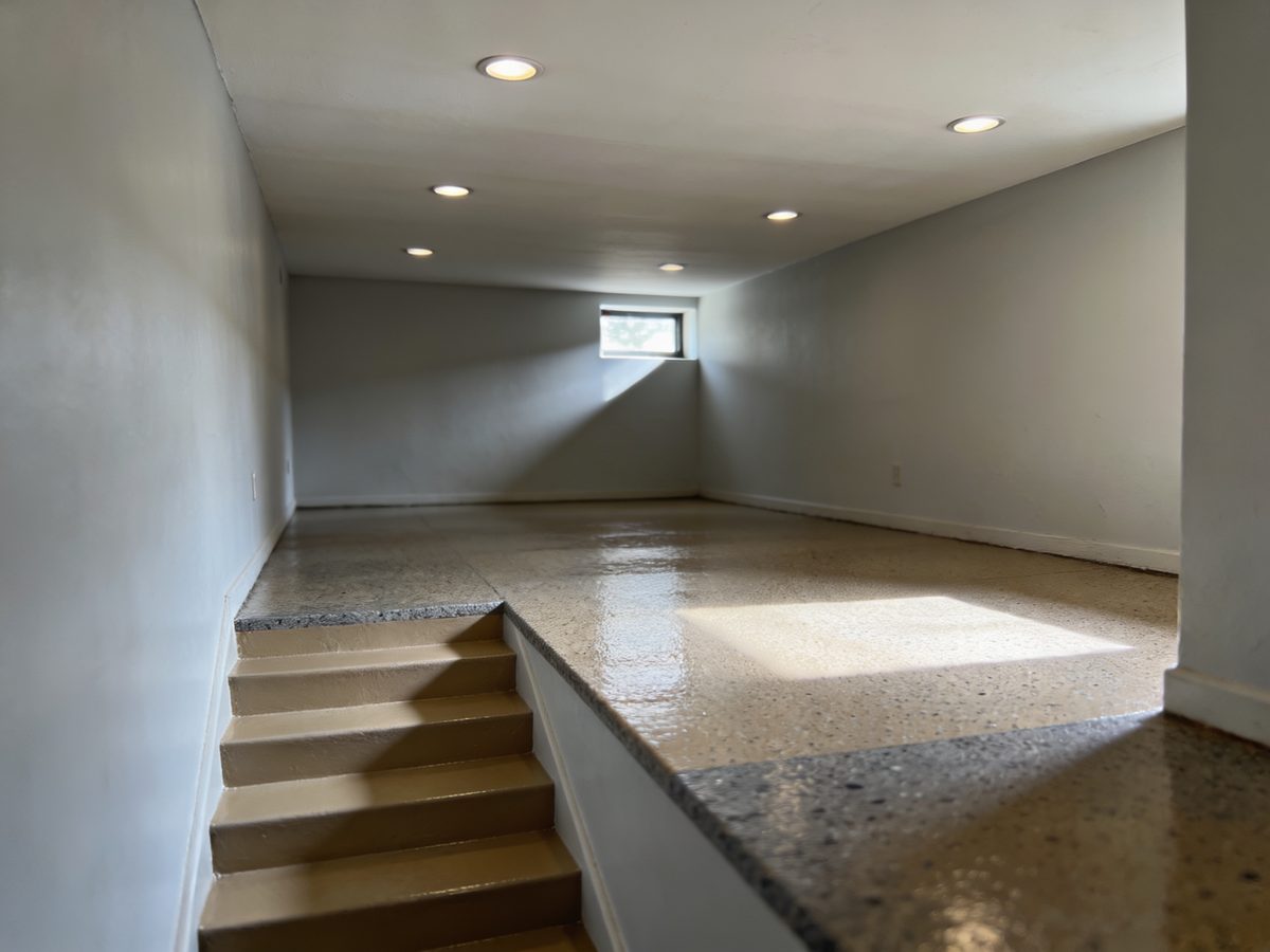 A basement rec room with a tan and slate flake floor coating, a painted stair and a window well throwing daylight across the floor